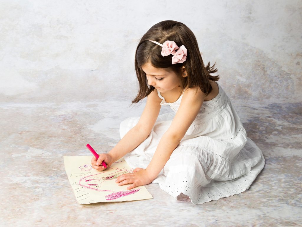 Niña con vestido blanco y lazo en el pelo dibuja sentada en el suelo.